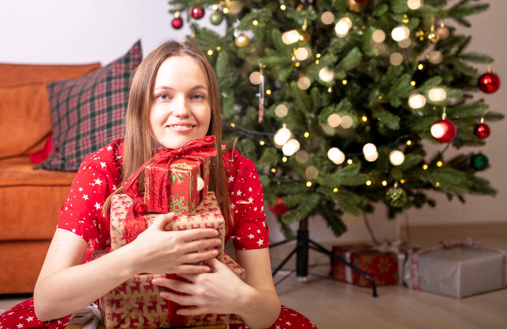 Młoda kobieta siedząca ze stosem opakowanych pudełek z prezentami w pobliżu choinki. Sam w domu z prezentami - Zdjęcie, obraz