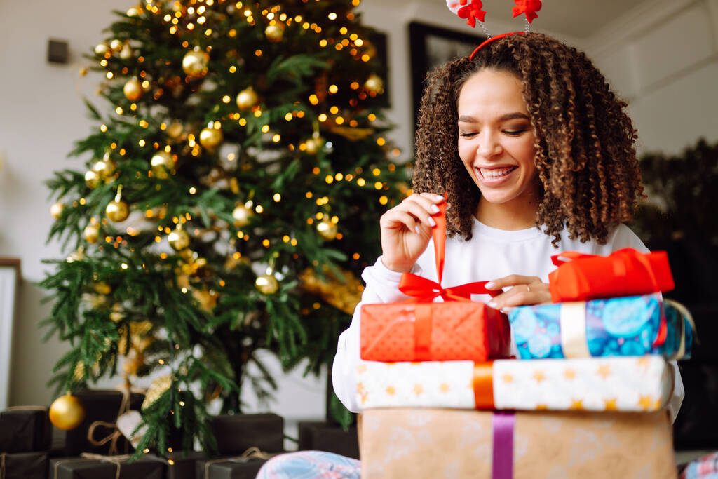 Onnellinen nainen avaa lahjoja lähellä joulukuusi. Nuori kihara nainen poseeraa lahjoja joulun sisustus. Muoti, juhla, loma käsite. - Valokuva, kuva