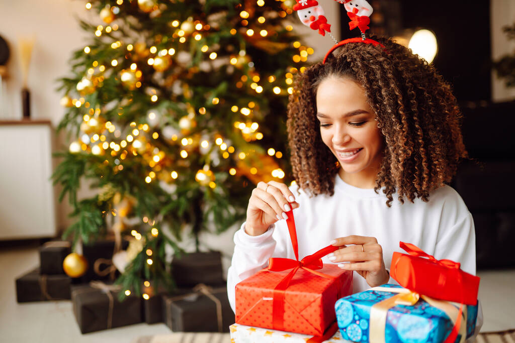 Ευτυχισμένη γυναίκα ανοίγει δώρα κοντά στο χριστουγεννιάτικο δέντρο. Νεαρή σγουρή κυρία ποζάρει με δώρα σε χριστουγεννιάτικο εσωτερικό σχέδιο. Μόδα, γιορτή, διακοπές έννοια. - Φωτογραφία, εικόνα