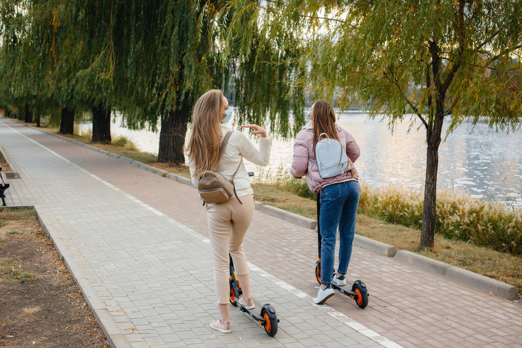 Dwie młode, piękne dziewczyny w maskach jeżdżą na skuterach elektrycznych w parku w ciepły jesienny dzień. Spacer po parku. - Zdjęcie, obraz