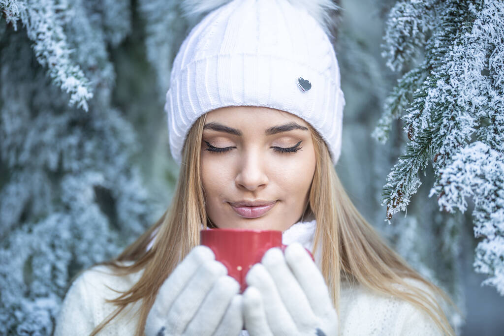 Fille blonde dans une forêt de conifères congelés sent boisson chaude dans une tasse rouge avec les yeux fermés, tous habillés en vêtements d'hiver blancs.  - Photo, image