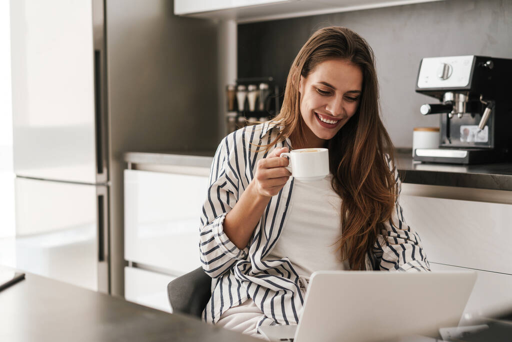 Nuori kaunis valkoihoinen nainen työskentelee kannettavan tietokoneen parissa ja juo kahvia kotona - Valokuva, kuva