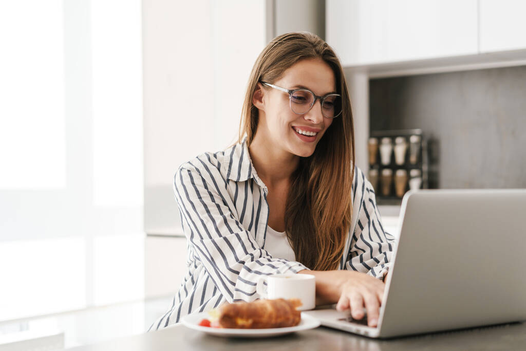 Nuori kaunis valkoihoinen nainen juo kahvia työskennellessään kannettavan tietokoneen kotona - Valokuva, kuva