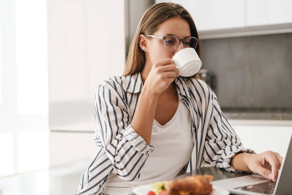 Νεαρή όμορφη καυκάσιος γυναίκα πίνοντας καφέ, ενώ εργάζονται σε φορητό υπολογιστή στο σπίτι - Φωτογραφία, εικόνα