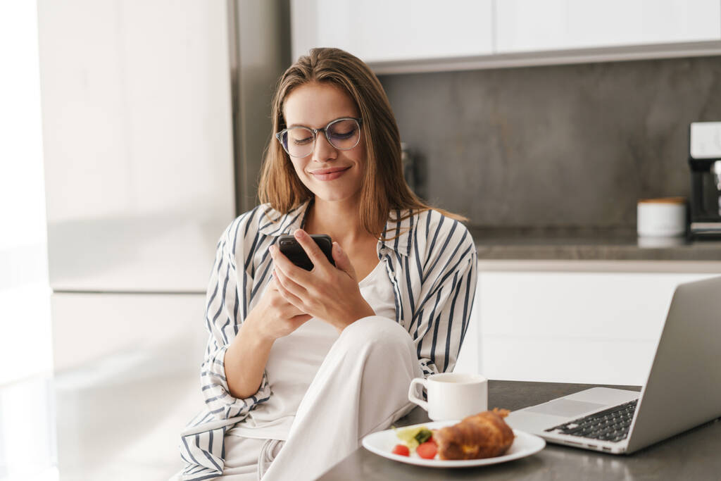 Joven hermosa mujer caucásica sosteniendo el teléfono celular mientras desayuna en casa - Foto, imagen
