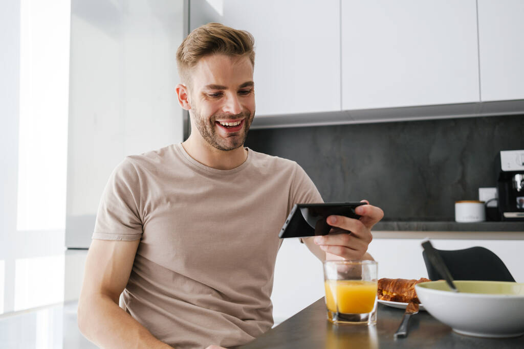 Mladý pohledný běloch drží mobilní telefon při snídani doma - Fotografie, Obrázek