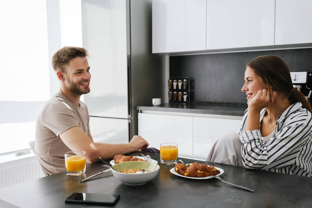 Jovem belo casal caucasiano sentado à mesa enquanto toma café da manhã em casa - Foto, Imagem