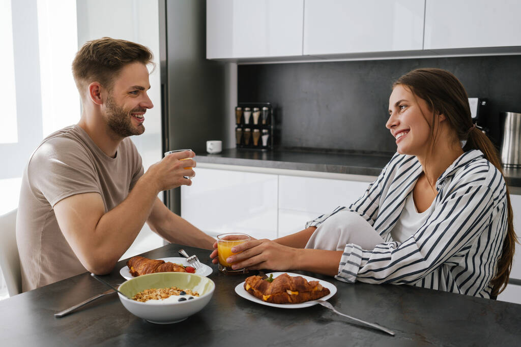 Junges schönes kaukasisches Paar sitzt am Tisch, während es zu Hause frühstückt - Foto, Bild