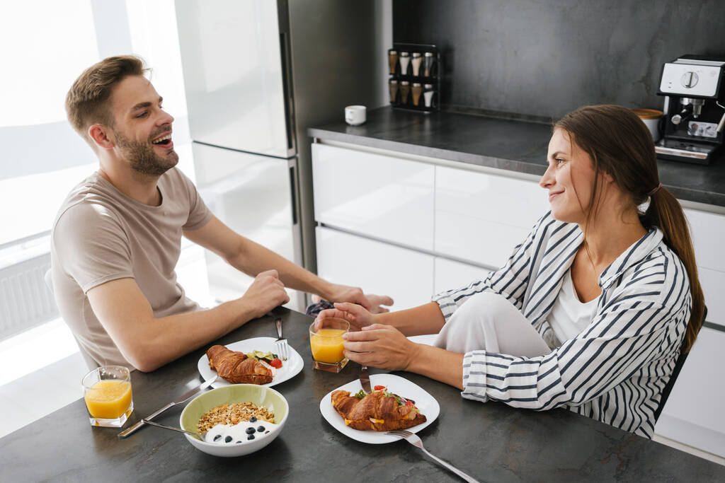 Jovem belo casal caucasiano sentado à mesa enquanto toma café da manhã em casa - Foto, Imagem
