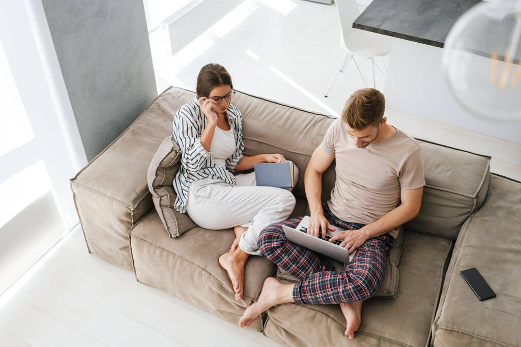 Jovem casal caucasiano segurando laptop e livro de leitura enquanto sentado no sofá em casa - Foto, Imagem