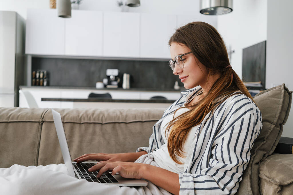 Fiatal kaukázusi boldog nő mosolyog és használja laptop, miközben ül a kanapén otthon - Fotó, kép
