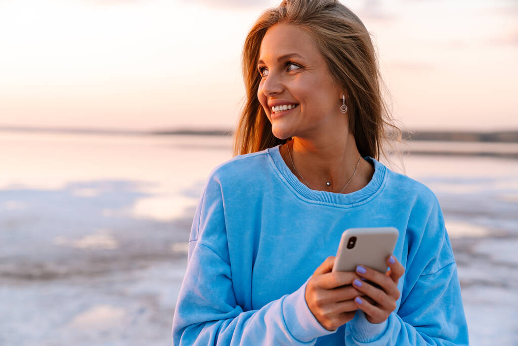 Jovem caucasiano feliz mulher loira sorrindo e segurando celular ao ar livre - Foto, Imagem