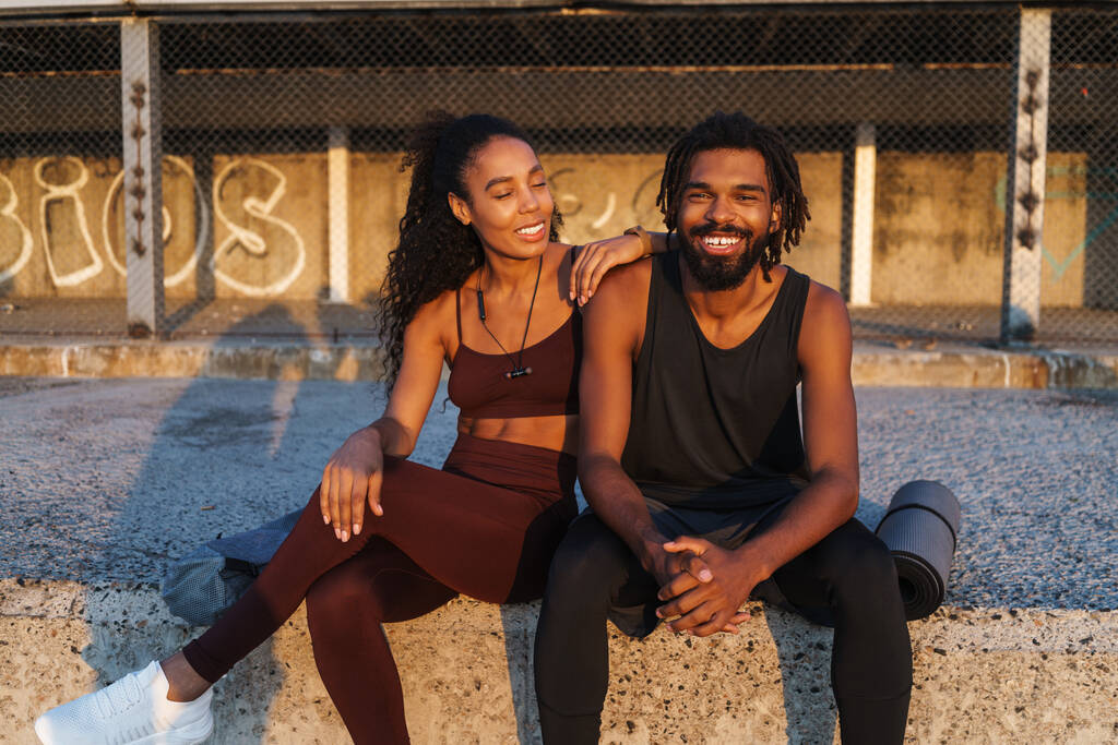 Молодий афроамериканець і жінка в костюмах посміхаються, сидячи надворі з матою для фізичних вправ. - Фото, зображення