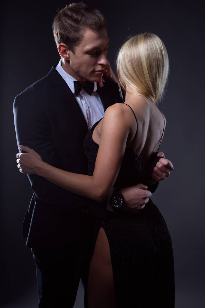 Passionate couple: a woman with a light hairstyle in a black evening dress and a handsome man in a suit with a bow tie pose in a dark Studio - Photo, Image