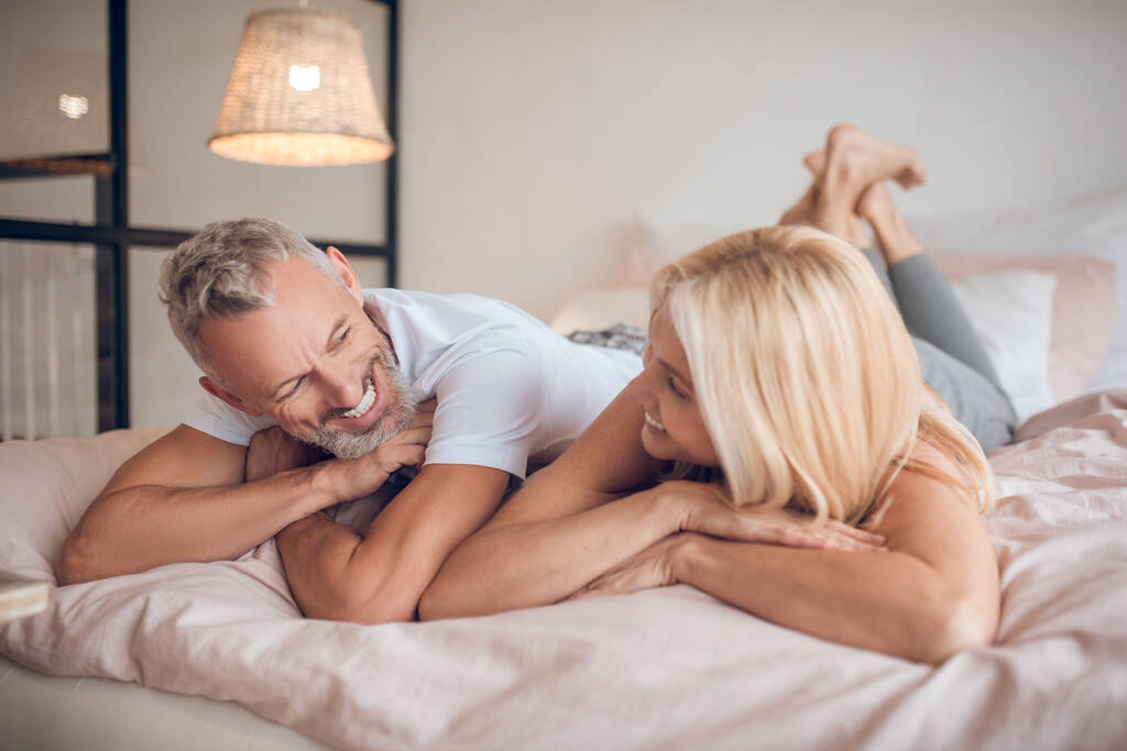 Седовласый мужчина и блондинка лежат на кровати и смотрят друг на друга. - Фото, изображение