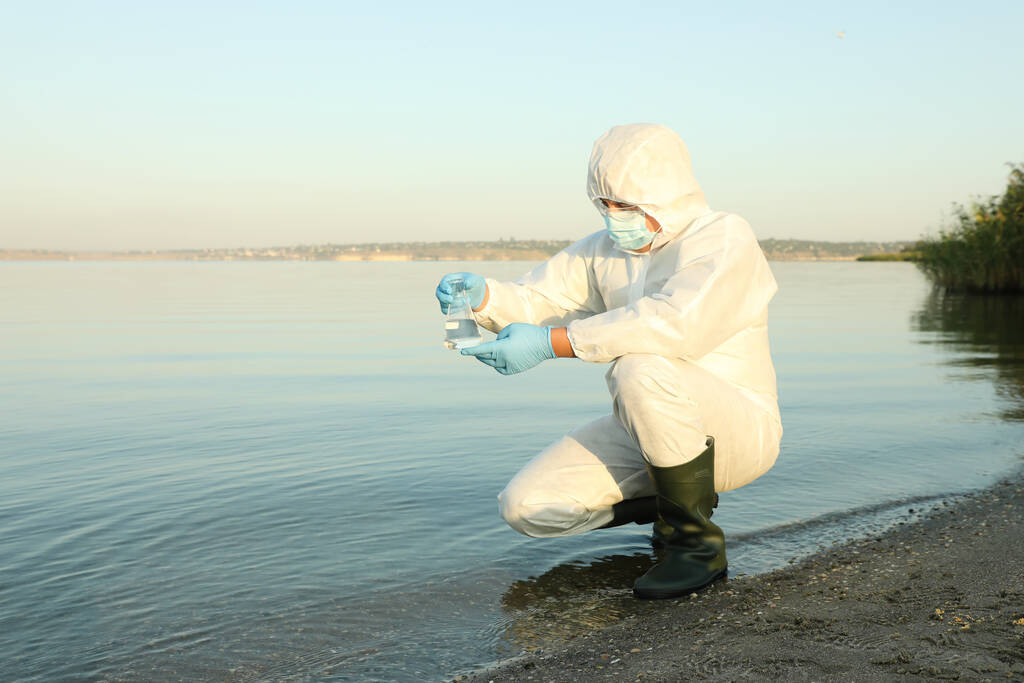 Wissenschaftler im Chemikalienschutzanzug mit konischem Kolben entnimmt Probe aus Fluss zur Analyse - Foto, Bild