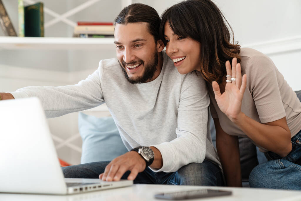 Image of cute cheerful couple using laptop and waving hand while sitting on sofa at home - Photo, Image