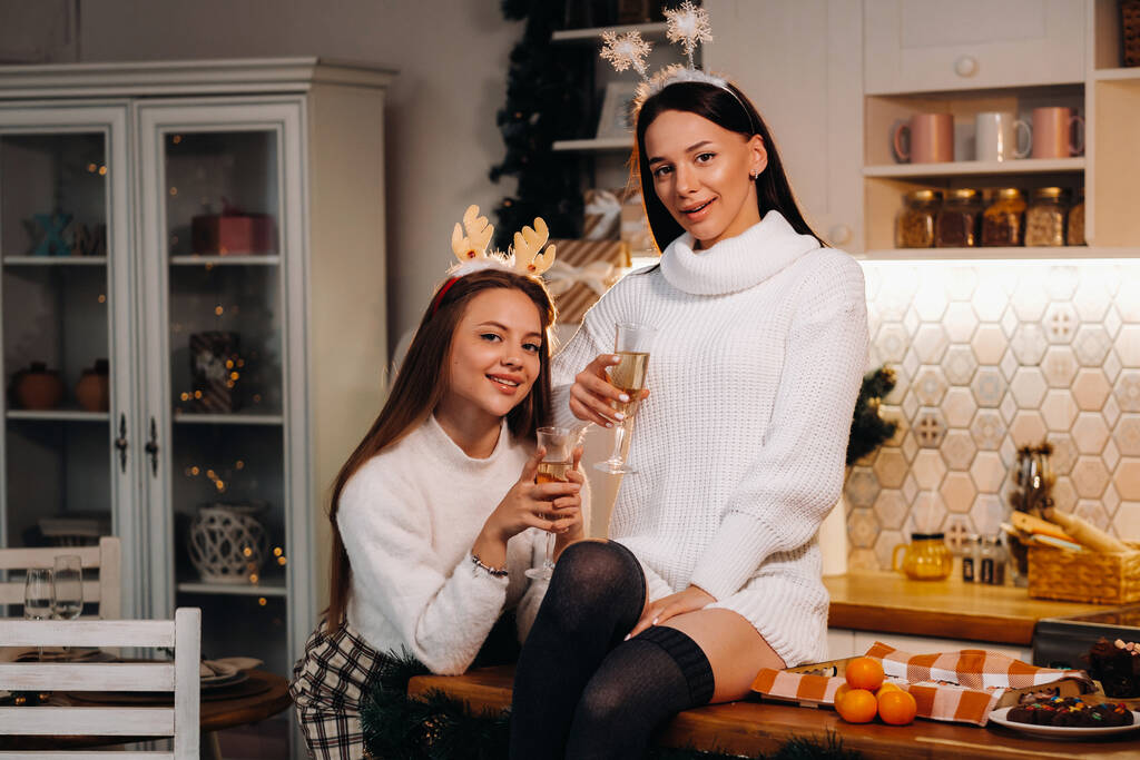 kaksi tyttöä viihtyisässä kotiympäristössä keittiössä samppanja käsissään jouluksi. Hymyilevät tytöt juovat samppanjaa juhlailtana.. - Valokuva, kuva