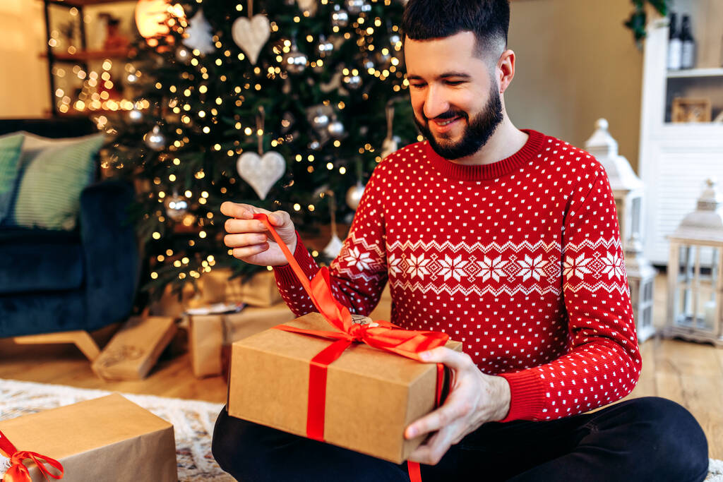 lächelnder junger Mann, der neben dem Weihnachtsbaum sitzt, mit einem Weihnachtsgeschenk zu Hause, Neujahrsfeier, frohe Weihnachten - Foto, Bild