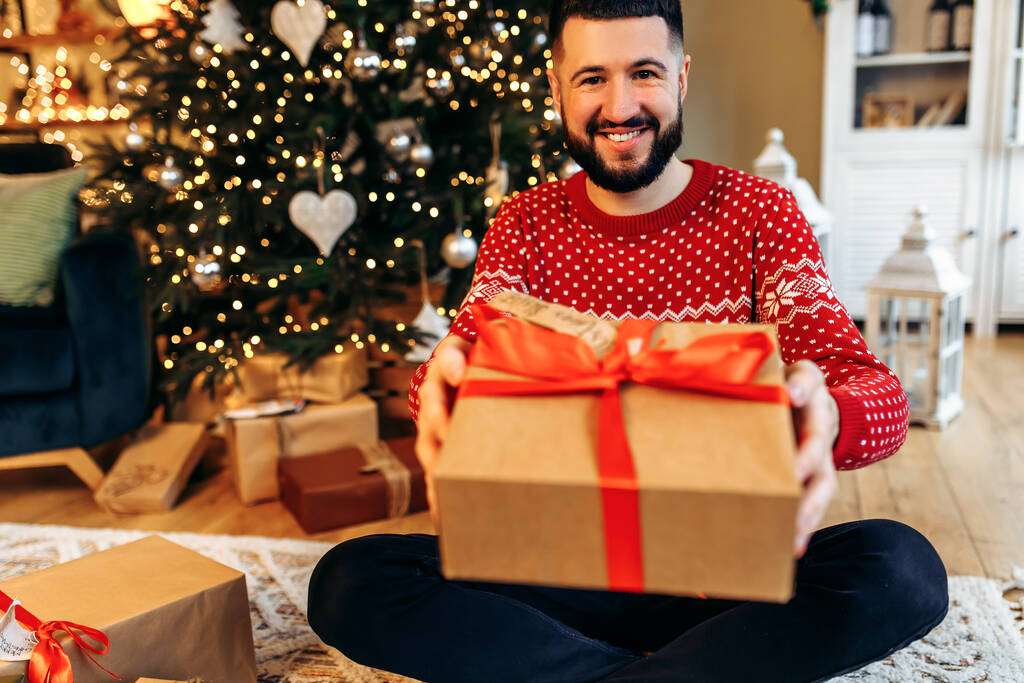 Smiling young man sitting near the Christmas tree, with a Christmas present at home, New Years celebration, Merry Christmas - Fotoğraf, Görsel