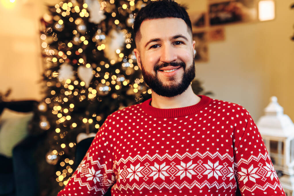 ein attraktiver junger Mann mit Bart, in rotem Pullover, sitzt zu Hause auf der Couch neben einem schönen, mit Spielzeug und Girlanden geschmückten Neujahrsbaum, frohe Weihnachten und Neujahr - Foto, Bild