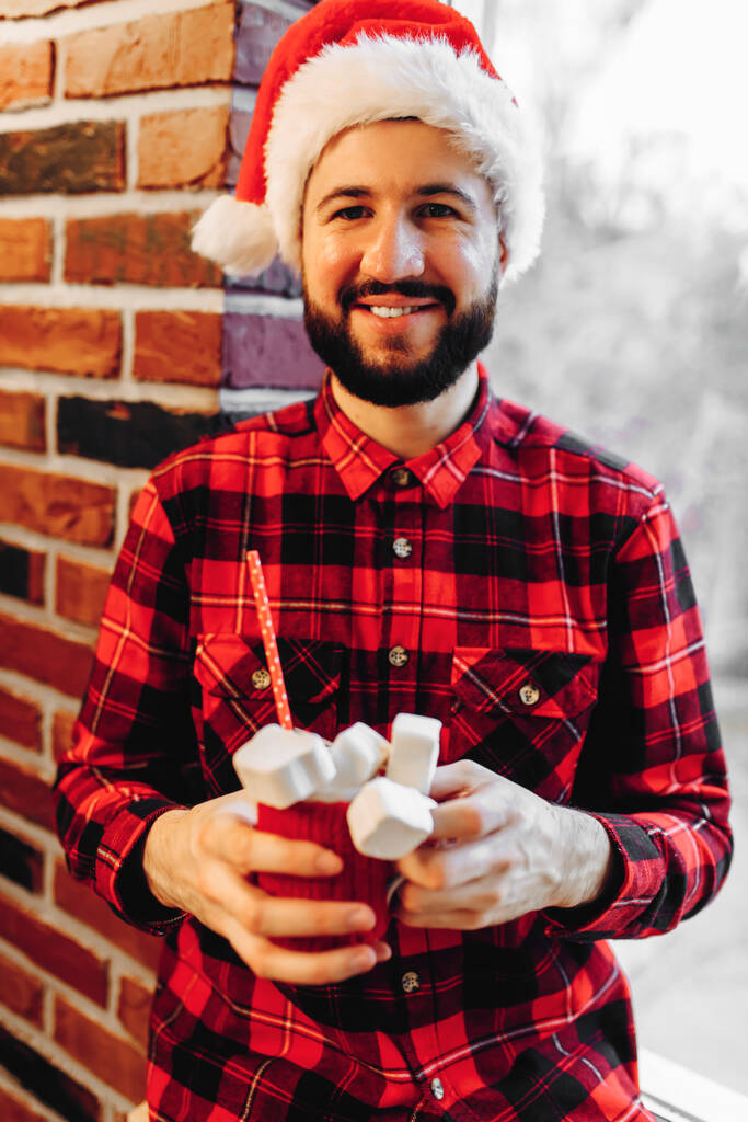 Portrait of a happy man in a Santa Claus hat, with a cup of coffee in his hands on a brick wall background, celebrating Christmas at home - Valokuva, kuva