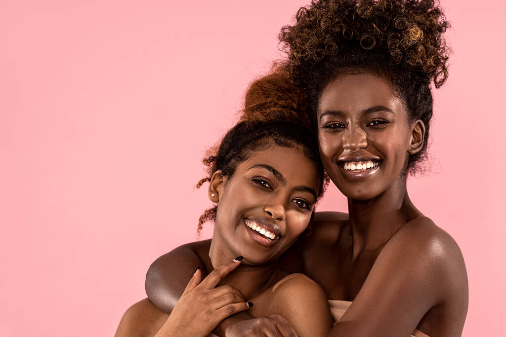 Birlikte poz veren, gülümseyen ve kameraya bakan heyecanlı genç kadınlar. Pembe pastel stüdyo arka planı. İki afro kadın modelin güzel portresi. Mükemmel dişli gülümsemeler.. - Fotoğraf, Görsel
