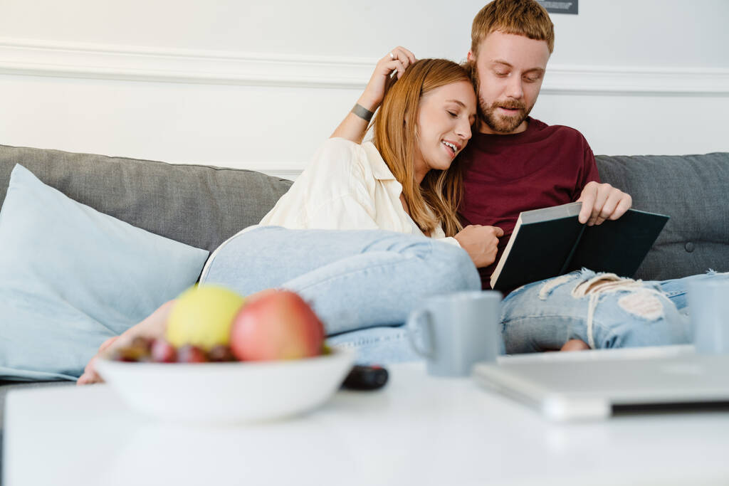 Photo of happy romantic couple hugging and reading book while sitting on couch at living room - Photo, Image