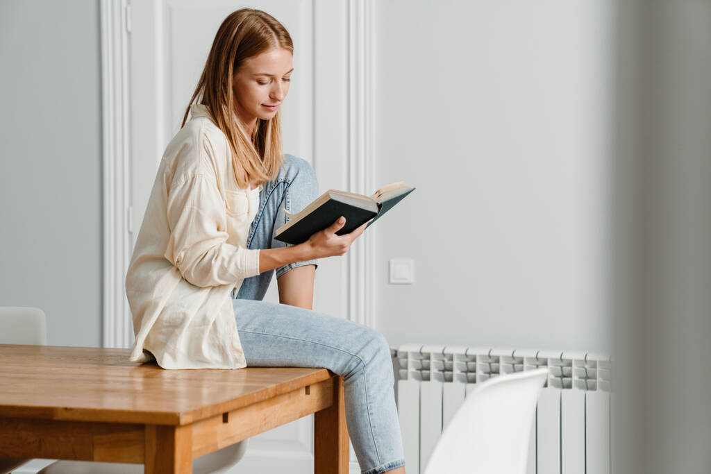 Afbeelding van gember gericht meisje lezen boek terwijl zitten op tafel thuis - Foto, afbeelding