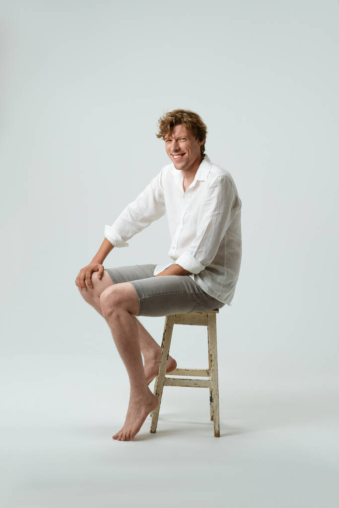 Volledige lengte van een zitten op stoel lachende jongeman kijken naar camera geïsoleerd op witte achtergrond. Gezichtsuitdrukkingen, emoties, gevoelens - Foto, afbeelding