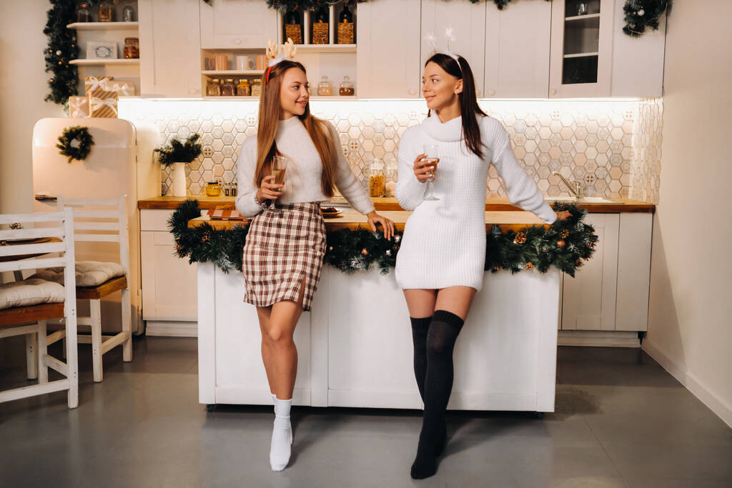 Két lány egy otthonos környezetben pezsgővel a kezében karácsonykor. Mosolygó lányok pezsgőt isznak egy ünnepi estén.. - Fotó, kép