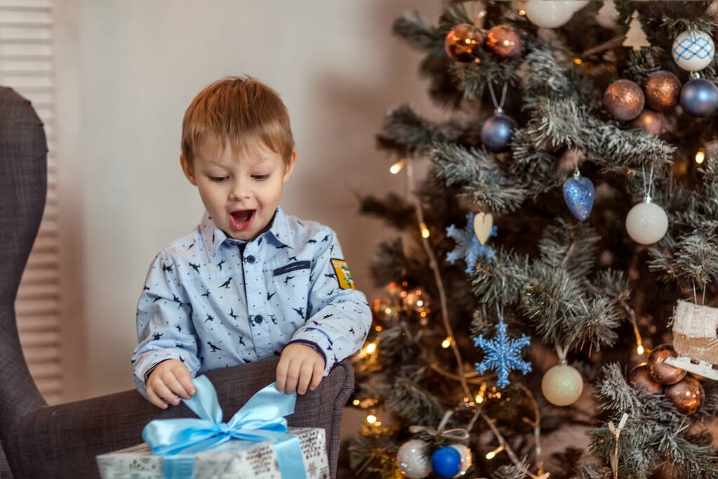 Kaunis 4-5-vuotias poika seisoo sohvan lähellä ja pitää lahjoja käsissään. Käsite talvilomat. Selektiivinen pehmeä fokus, bokeh (hämärtynyt tausta). - Valokuva, kuva