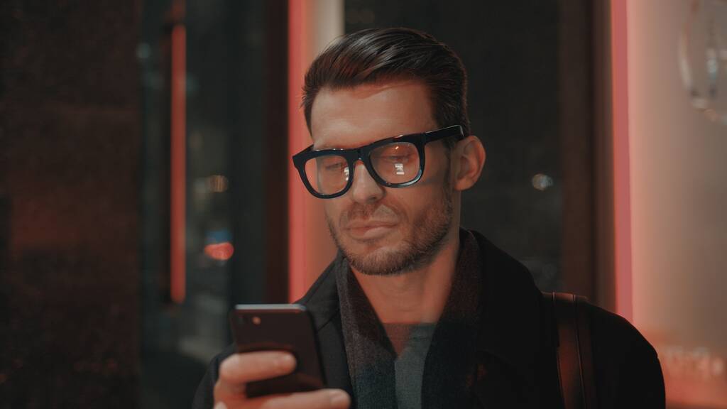 Mies silmälaseissa puhelimen kanssa liikerakennuksen taustalla yöllä - Valokuva, kuva