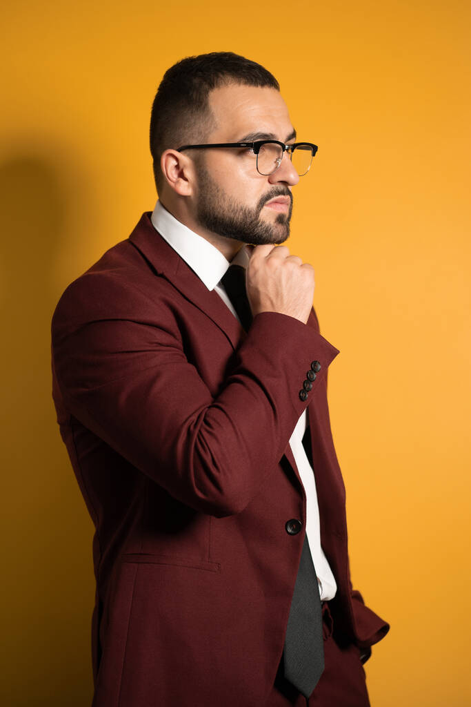 Handsome young man eye glasses wearing burgundy suit looking seriously away with hand folded touching chin standing sideways isolated on yellow background - Photo, Image