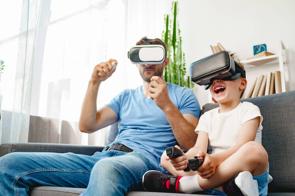 Baba oğul kanepede oturup VR gözlükleriyle oyun oynuyorlar. - Fotoğraf, Görsel