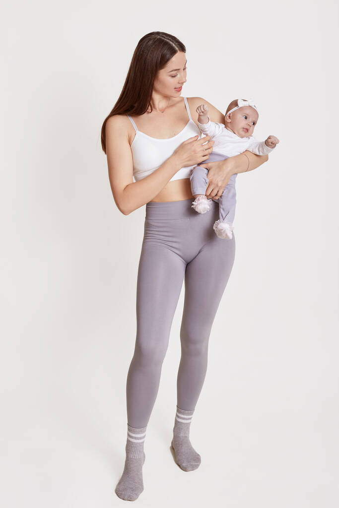 Full length portret van moeder staan en houden baby in handen, dame dragen beha en grijs leggins, poseren geïsoleerd over witte achtergrond, mama spelen met baby meisje. - Foto, afbeelding