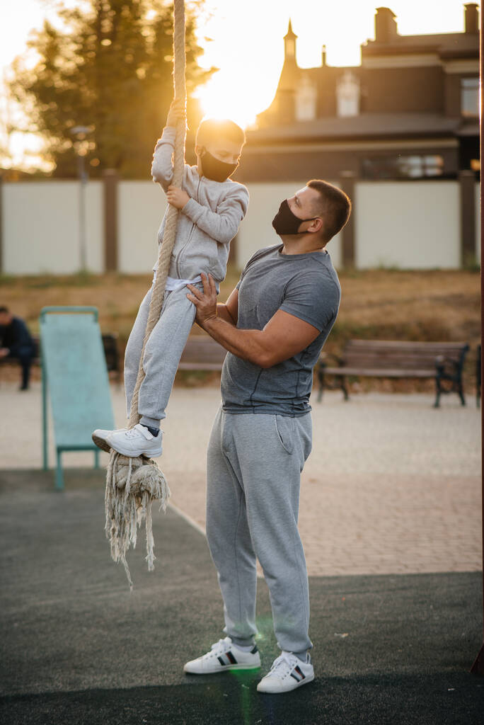 Батько допомагає синові піднятися на мотузку на спортивному полі в масках під час заходу сонця. Здорове виховання та здоровий спосіб життя
 - Фото, зображення