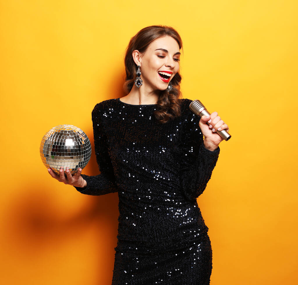 Concepto de fiesta, fiesta y celebración: Joven morena con el pelo largo rizado vestida con vestido de noche sosteniendo un micrófono y bola disco - Foto, Imagen