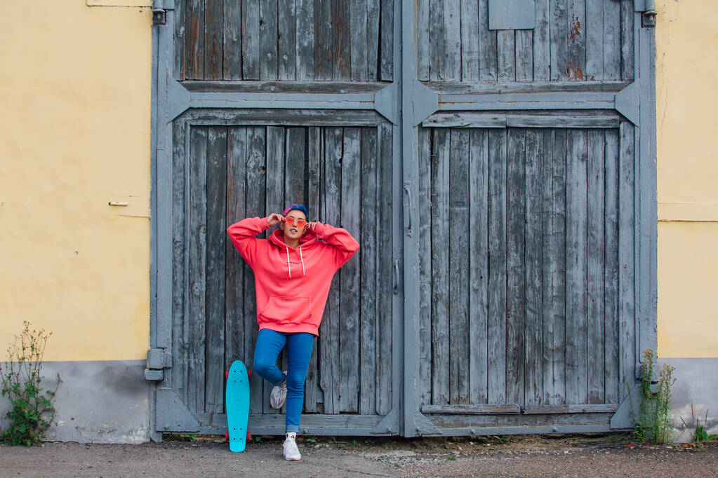 Stijlvolle jonge vrouw met kort gekleurd haar, gekleed in een roze hoodie en roze bril die bij oude houten poorten staat met haar plastic skateboard. Jeugdconcept. - Foto, afbeelding