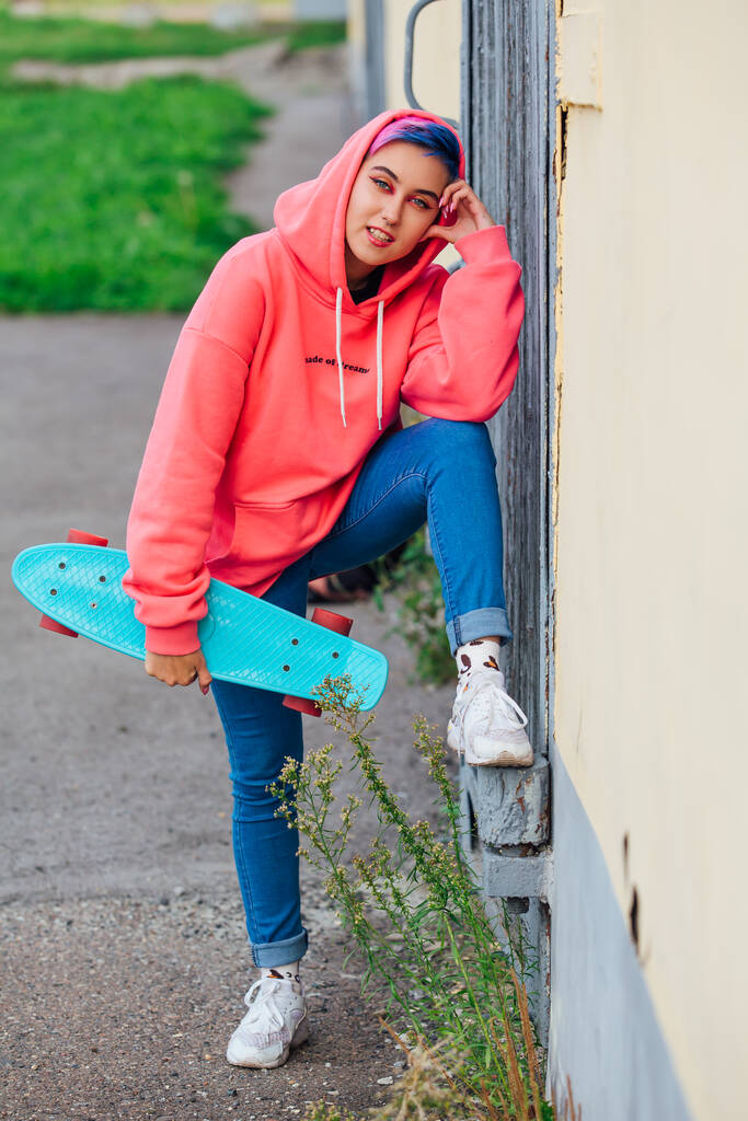 Κομψή νεαρή γυναίκα με κοντά χρωματιστά μαλλιά κρατώντας μπλε πλαστικό skateboard στο χέρι. Έννοια της νεολαίας. - Φωτογραφία, εικόνα