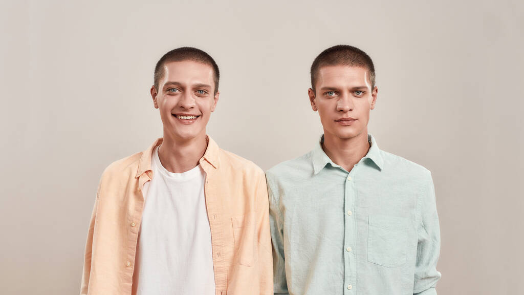 Portret dwóch młodych braci bliźniaków ubranych w luźne ubrania, patrzących przed kamerę i pozujących razem na beżowym tle. - Zdjęcie, obraz