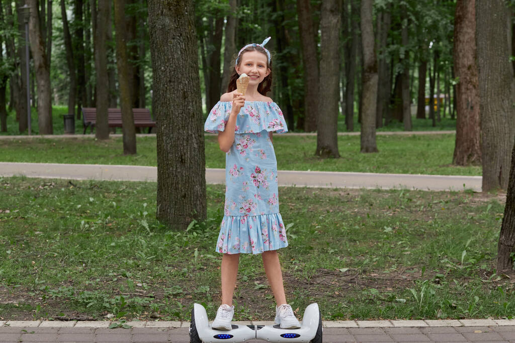 Une fille mange de la crème glacée douce, monte sur un scooter gyroscopique et regarde dans le cadre. - Photo, image