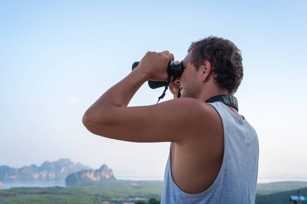 A young man in a white T-shirt looks through binoculars from the top against the background of the sky and rocks - Foto, afbeelding