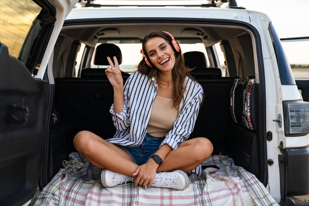 Молодая улыбающаяся молодая женщина в наушниках сидит на заднем сиденье автомобиля - Фото, изображение