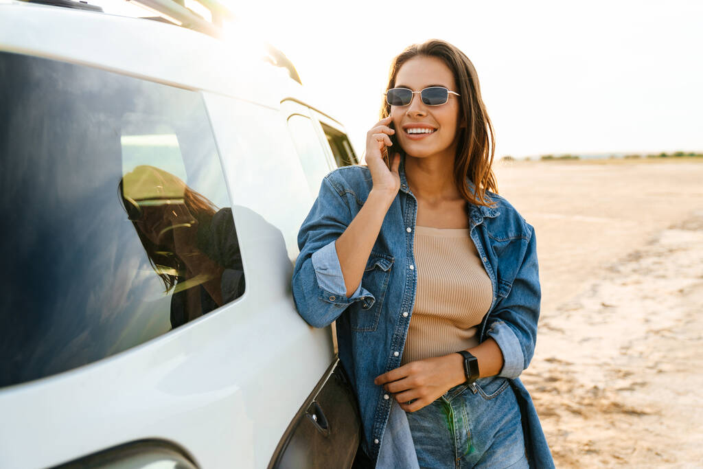Привлекательная молодая женщина, опирающаяся на автомобиль, стоя на пляже, используя мобильный телефон, разговаривая - Фото, изображение