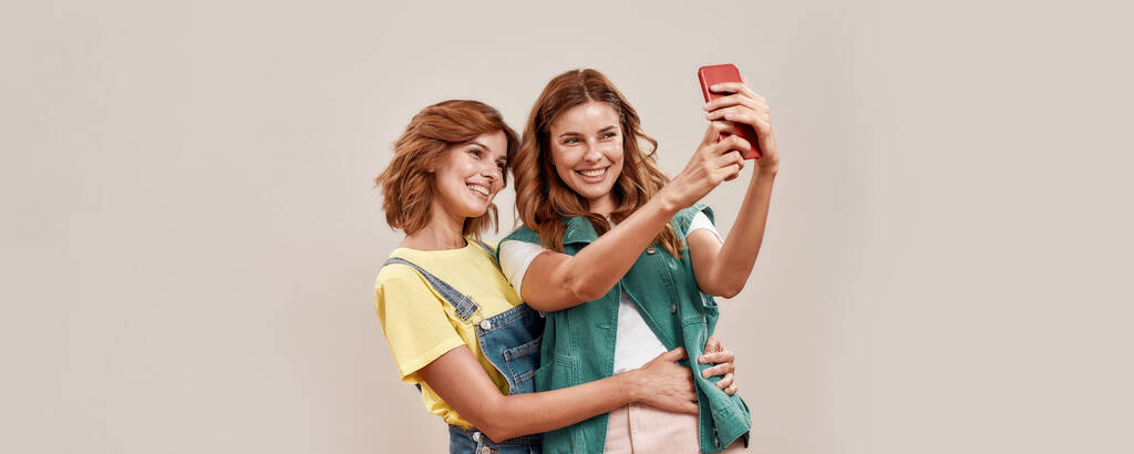 Due giovani ragazze attraenti, sorelle gemelle in abbigliamento casual sorridenti mentre si scattano selfie utilizzando il telefono cellulare, in piedi insieme isolati su sfondo chiaro - Foto, immagini