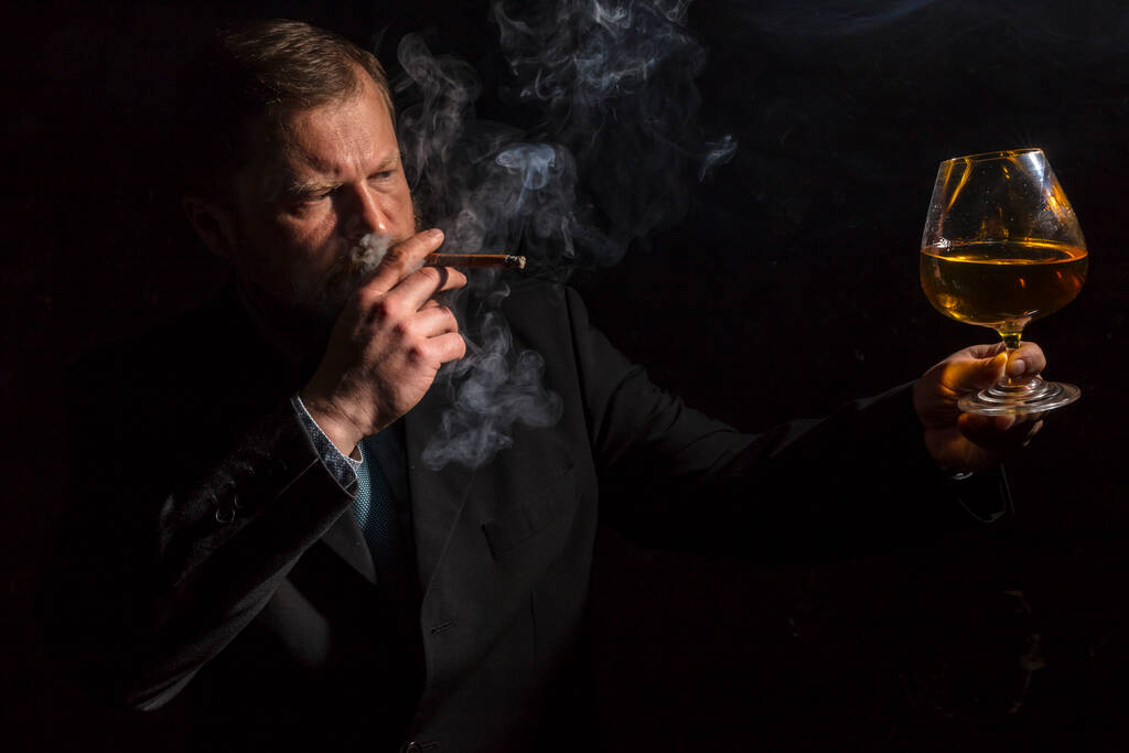 Твердолобый бородатый мужчина в костюме со стаканом виски и сигарой с дымом - Фото, изображение