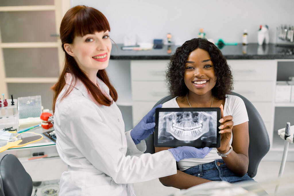 Prise de vue d'une dentiste professionnelle et d'une patiente africaine assise dans une chaise de dentisterie, montrant une tablette numérique avec balayage oral du patient, radiographie des mâchoires et des dents, à la caméra et souriant - Photo, image