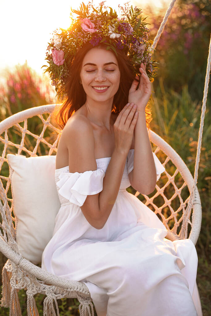 Gyönyörű virágkoszorút viselő fiatal nő hintaszékben a szabadban, naplementekor. - Fotó, kép