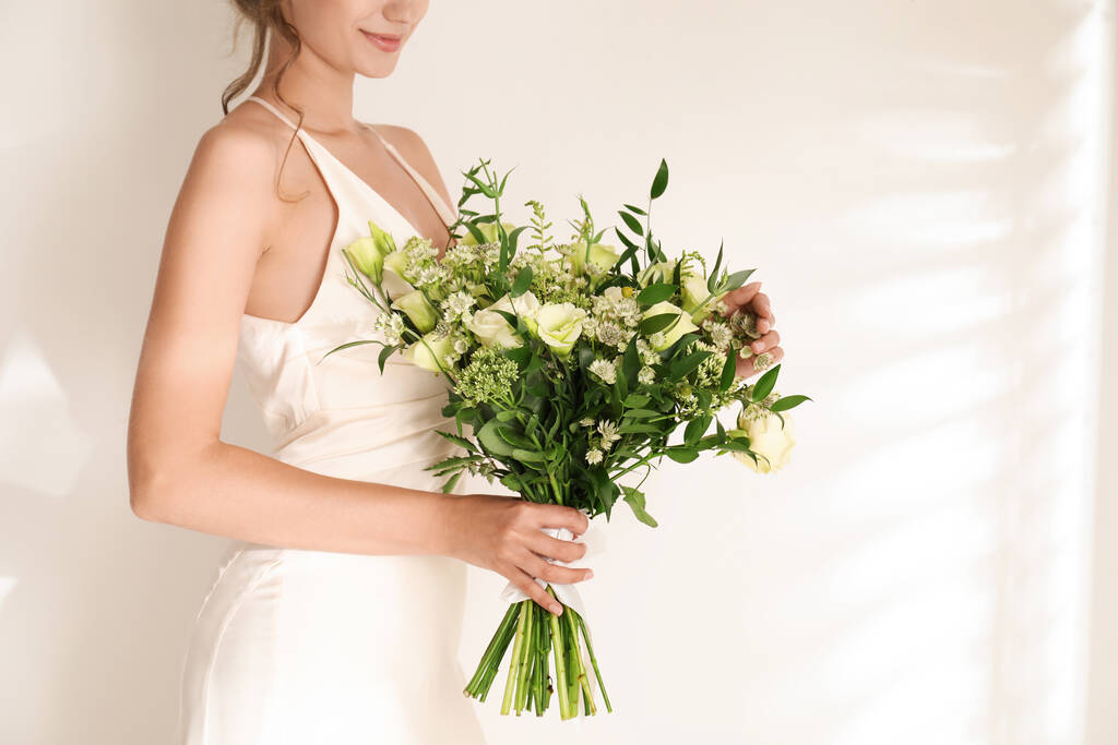 Νεαρή νύφη φορώντας νυφικό με όμορφο μπουκέτο στο φως φόντο, closeup - Φωτογραφία, εικόνα